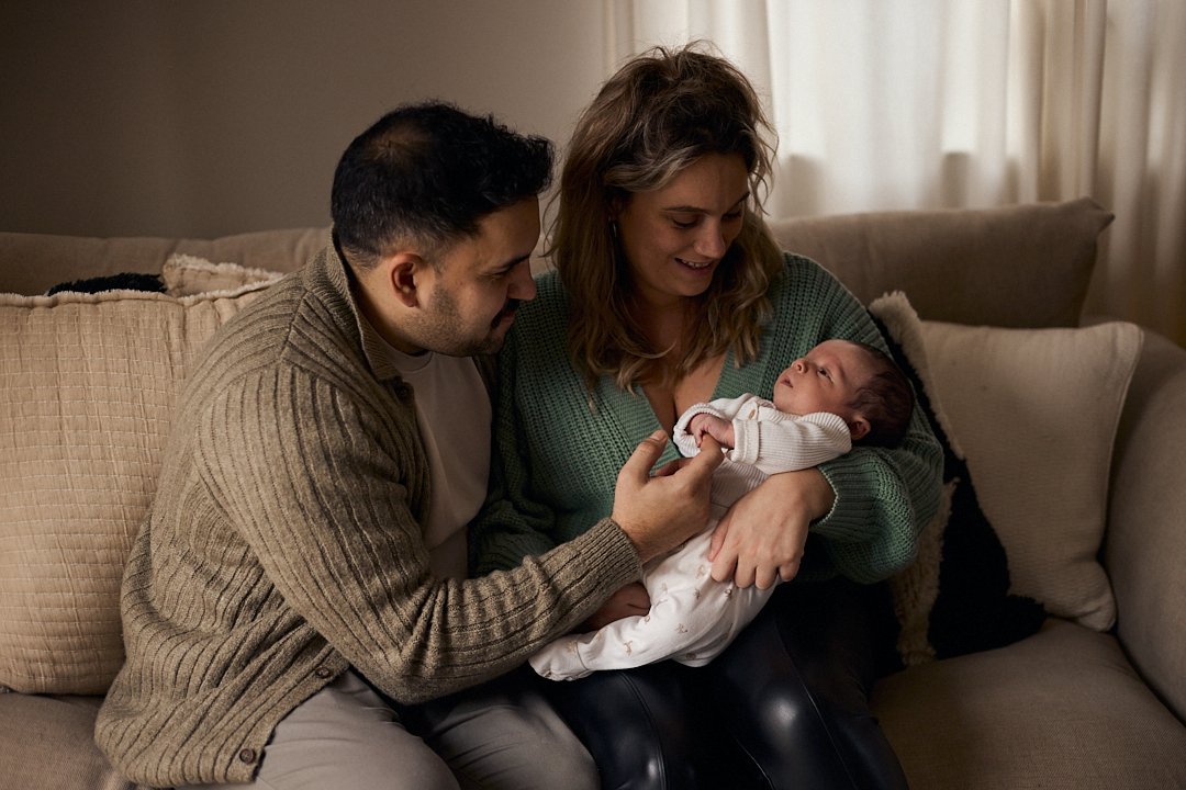 A smiling woman is sitting on a sofa holding a newborn baby in her arms, while a man leans in close, touching the baby's hand, all inside a cozy living room.