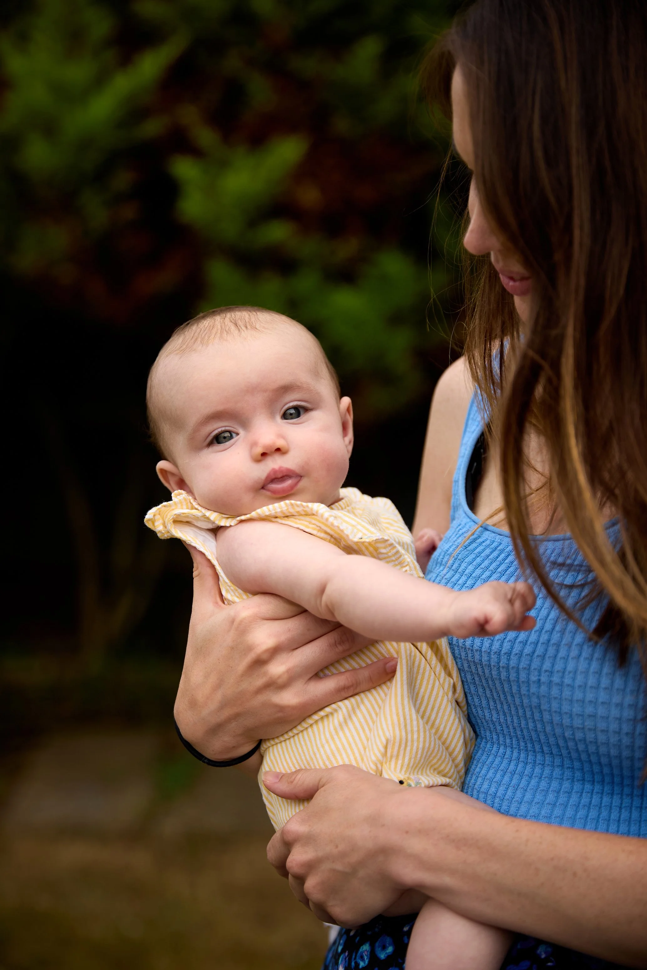 A woman holding a baby outdoors with green trees in the background.