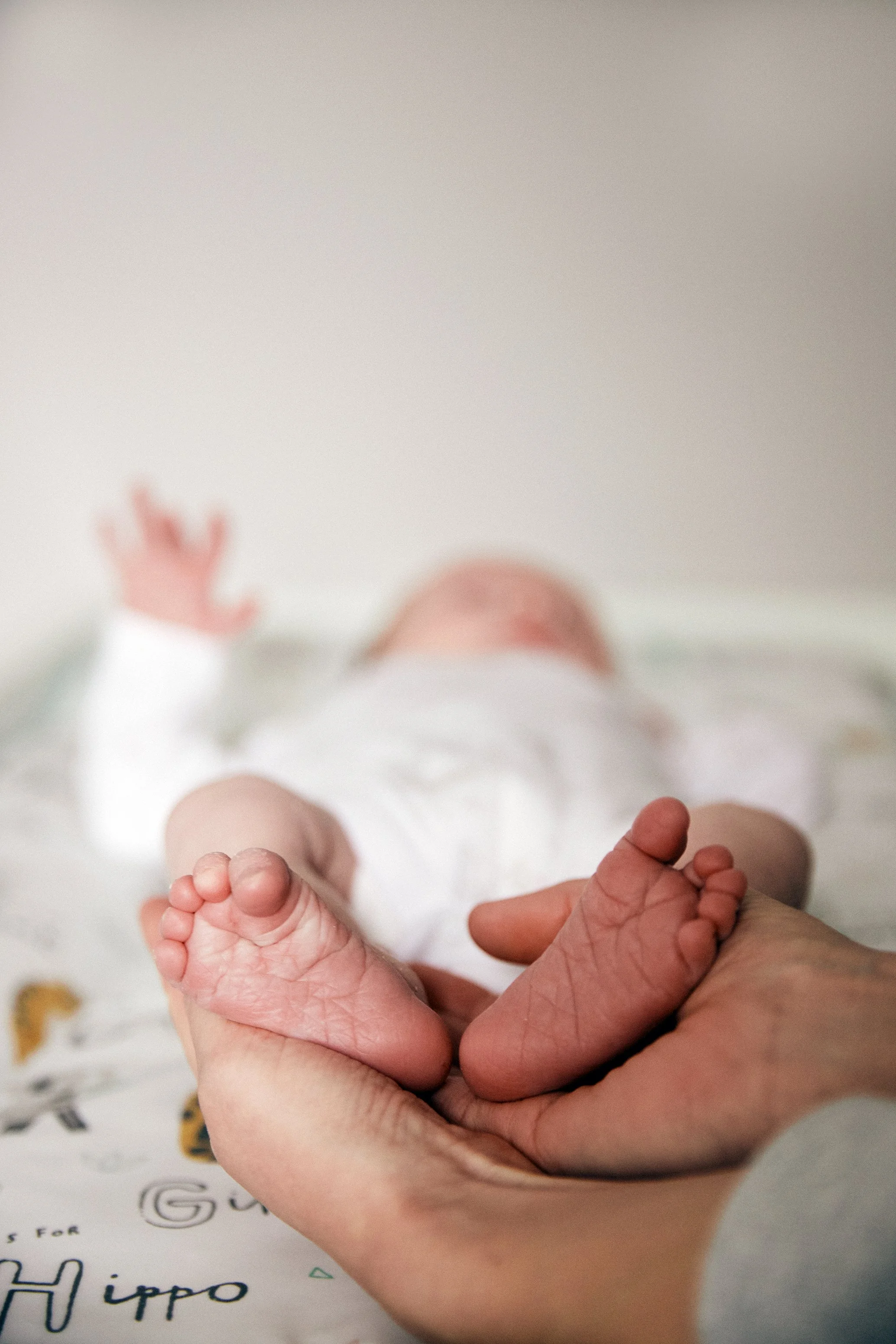 A newborn baby lying on a bed, being gently held by an adult's hand, with the baby's tiny feet and outstretched arm visible.