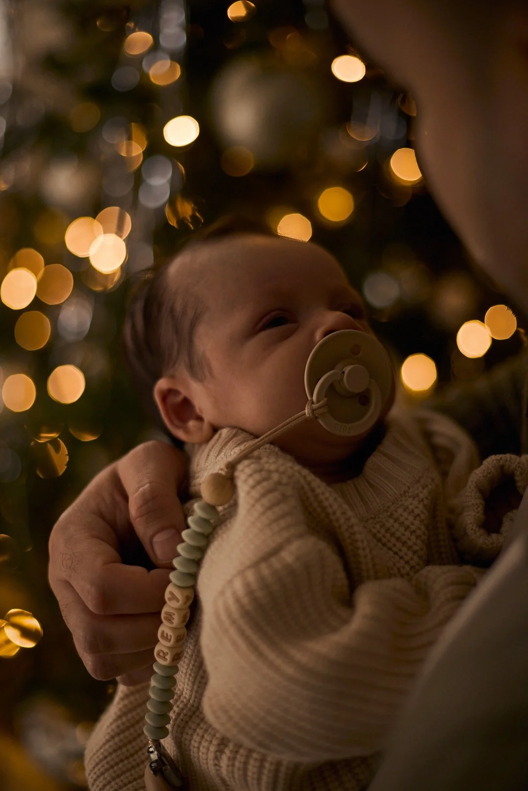 A newborn baby being held by an adult, looking up, with Christmas lights in the background.