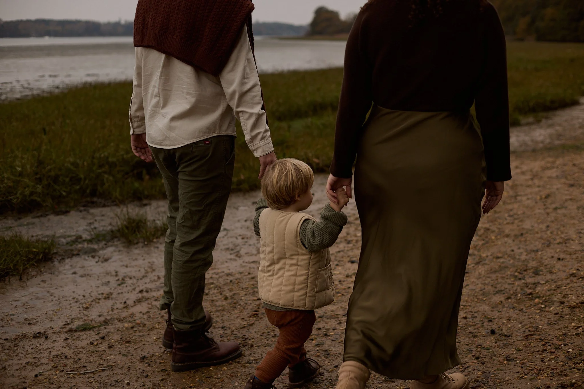 A family of three walking along a nature trail by a lake during sunset, holding hands.