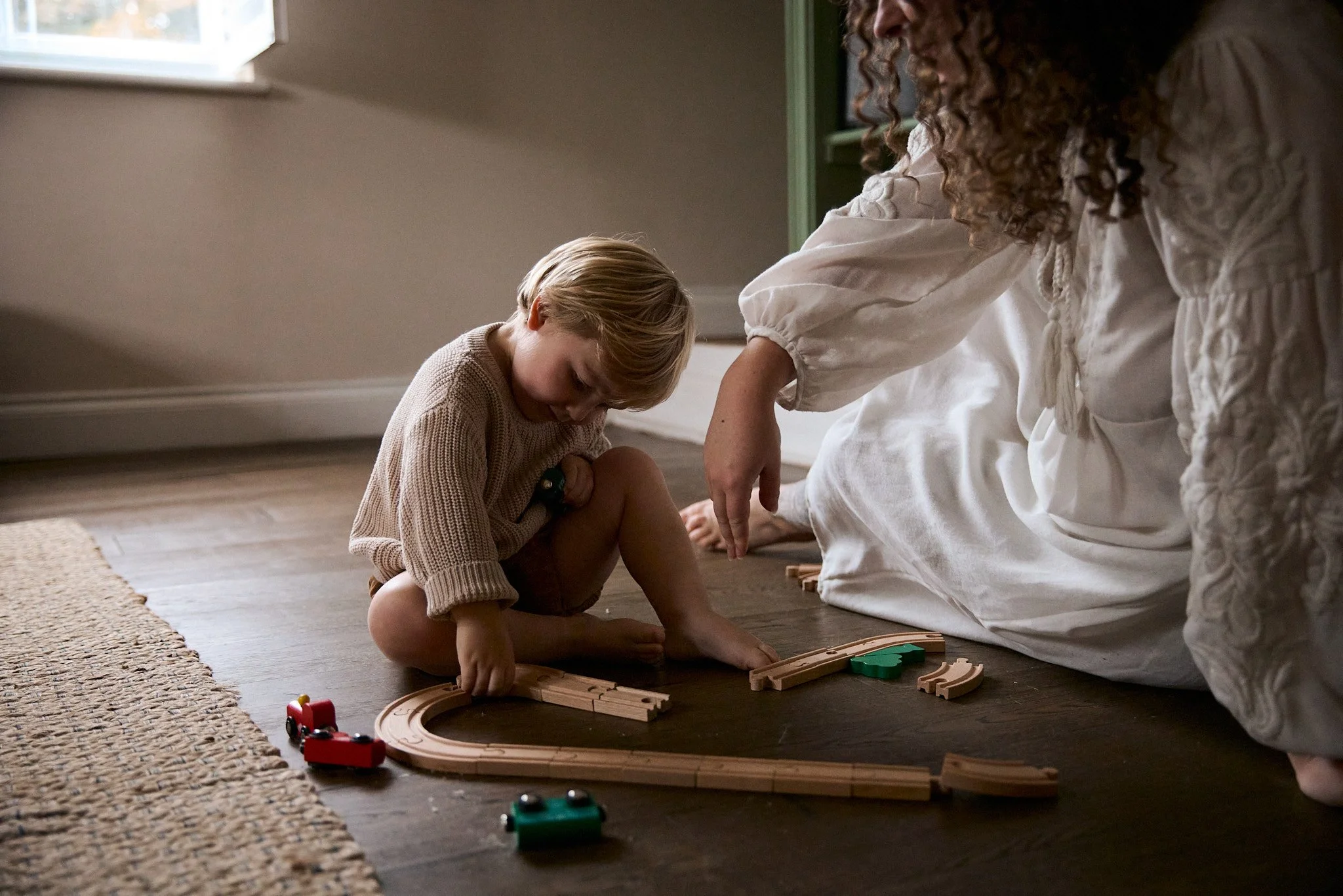 A young boy and a woman playing with wooden train tracks on a hardwood floor near a window, with a carpet partially visible on the side.