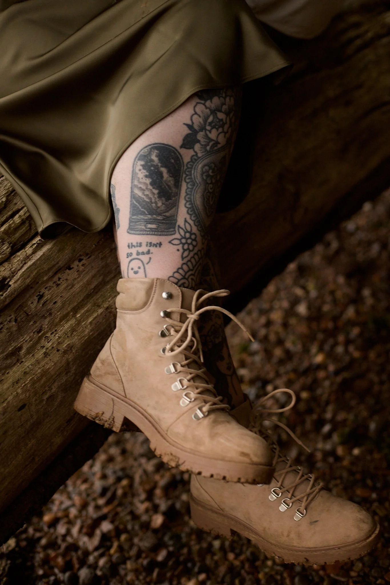Close-up of a person's bottom leg, showing tattoos, wearing beige combat boots, and khaki shorts, with a wooded log in the background.