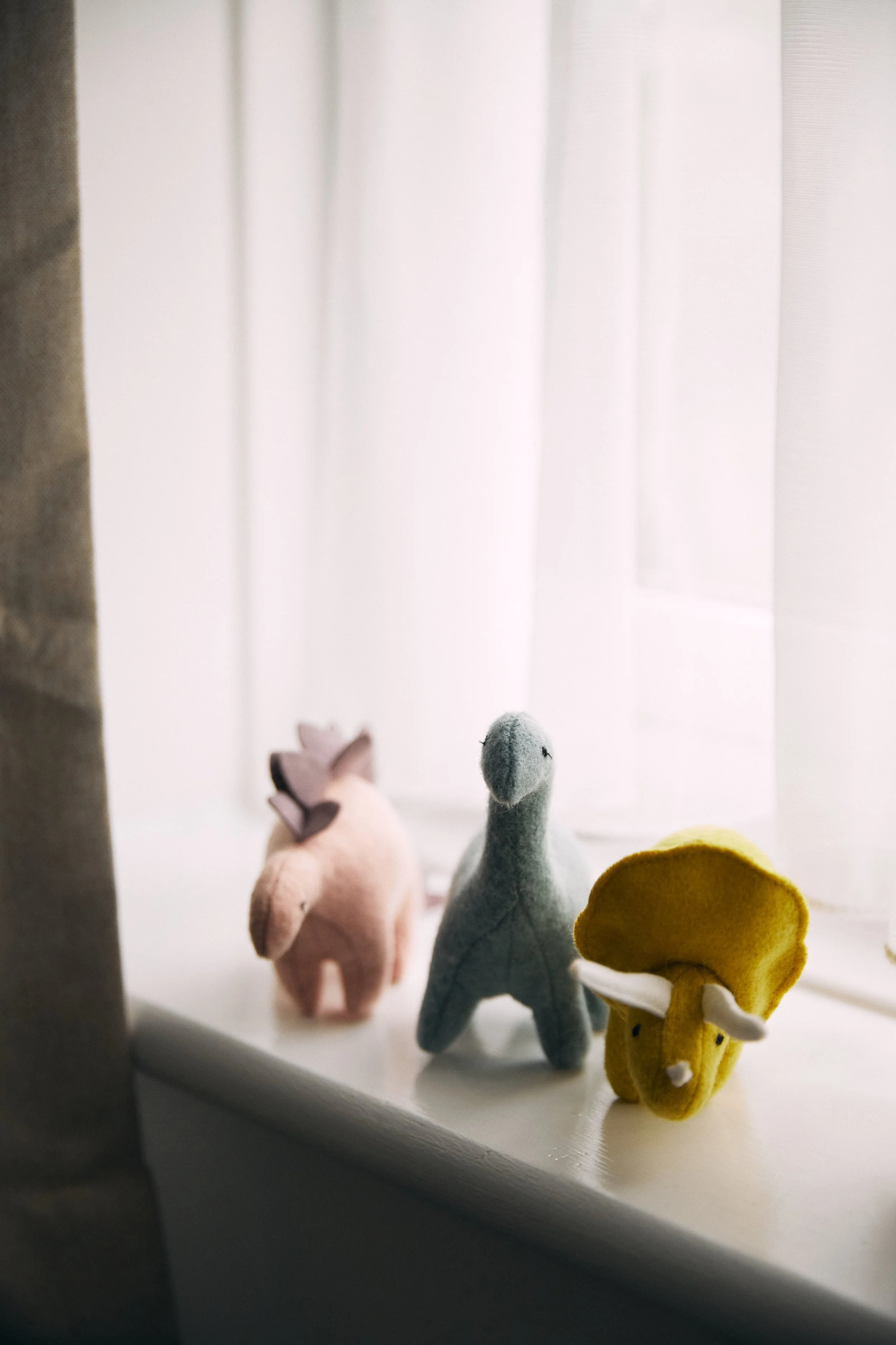 Three stuffed animals sit on a window sill: a pink pig, a gray dinosaur, and a yellow bison, with a sheer white curtain behind them.