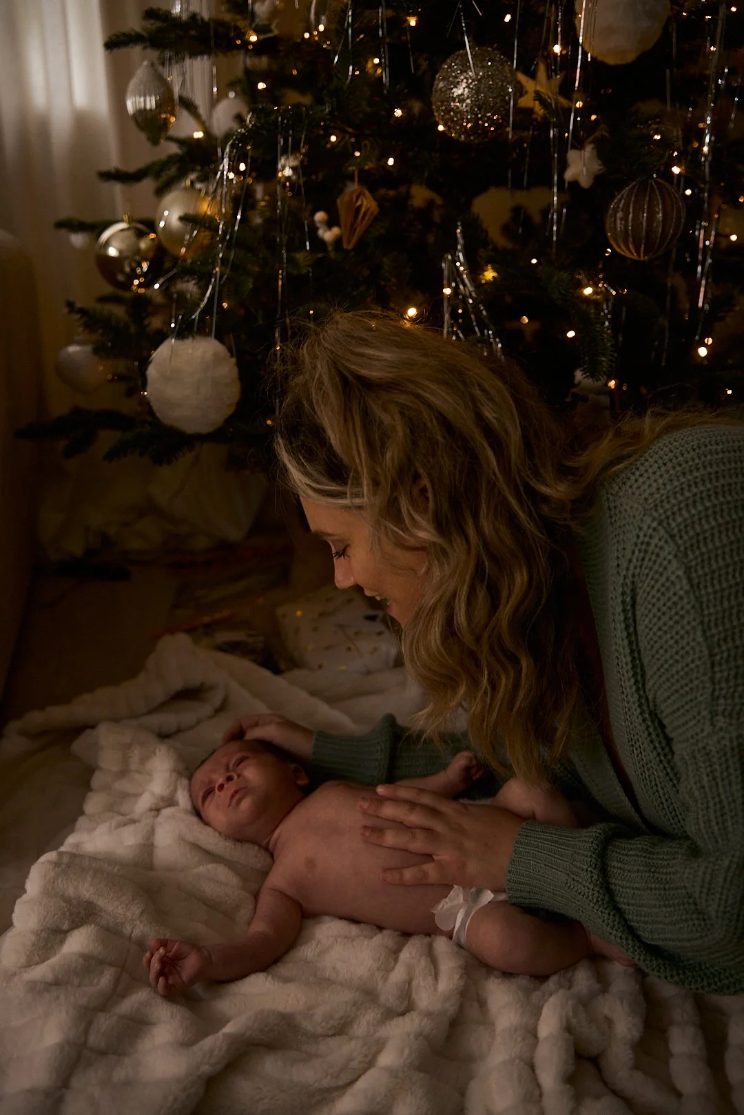 A woman with curly hair leaning over a young, shirtless child lying on a soft blanket near a Christmas tree decorated with ornaments and lights.