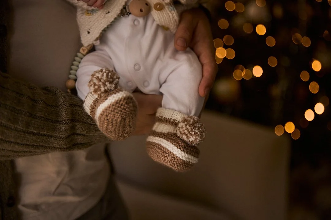 A baby being held by an adult, wearing cozy knit booties and a knitted sweater, with a Christmas tree with warm lights in the background.