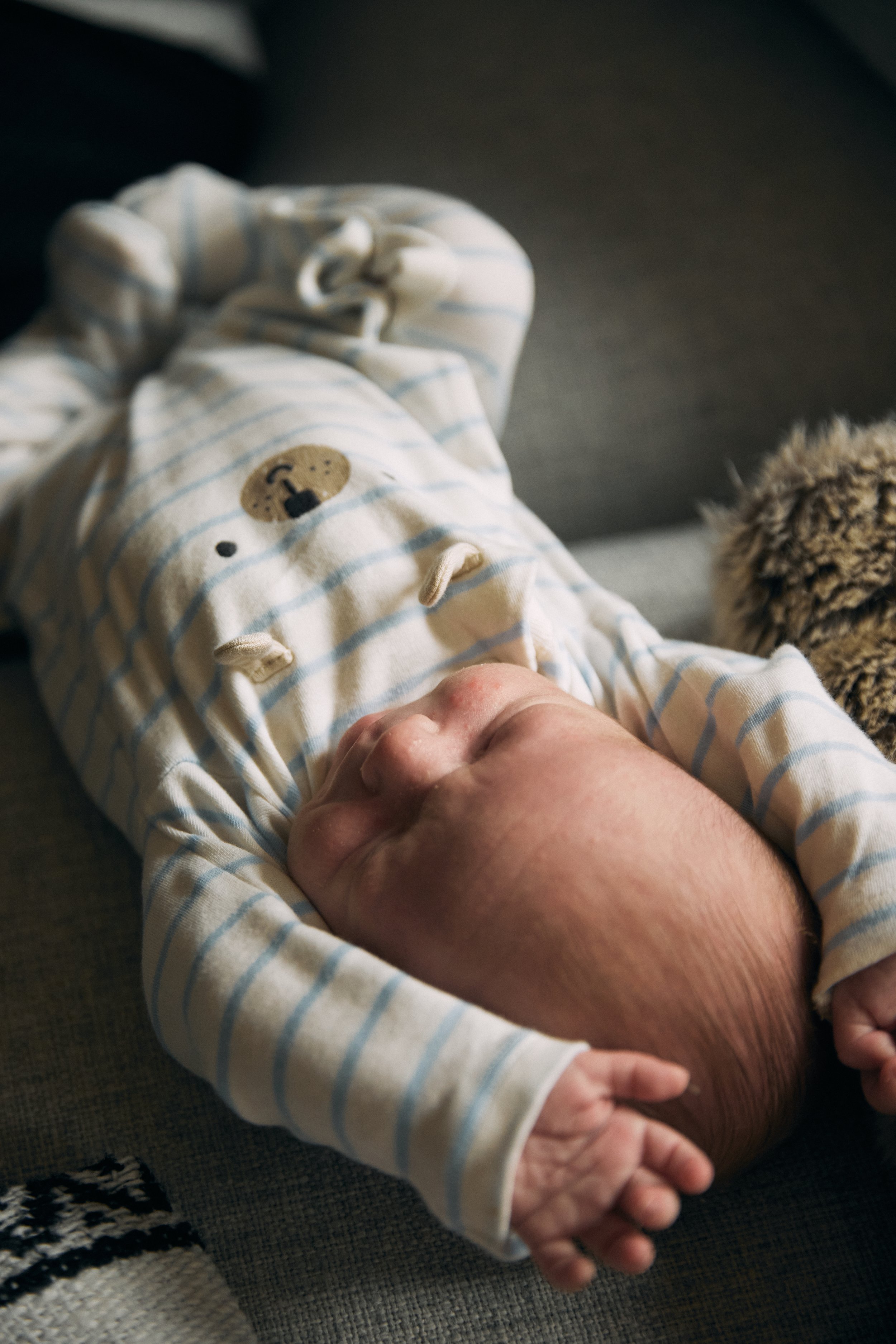 A sleeping baby lying on a dark couch, wearing striped pajamas with a bear face on the chest.