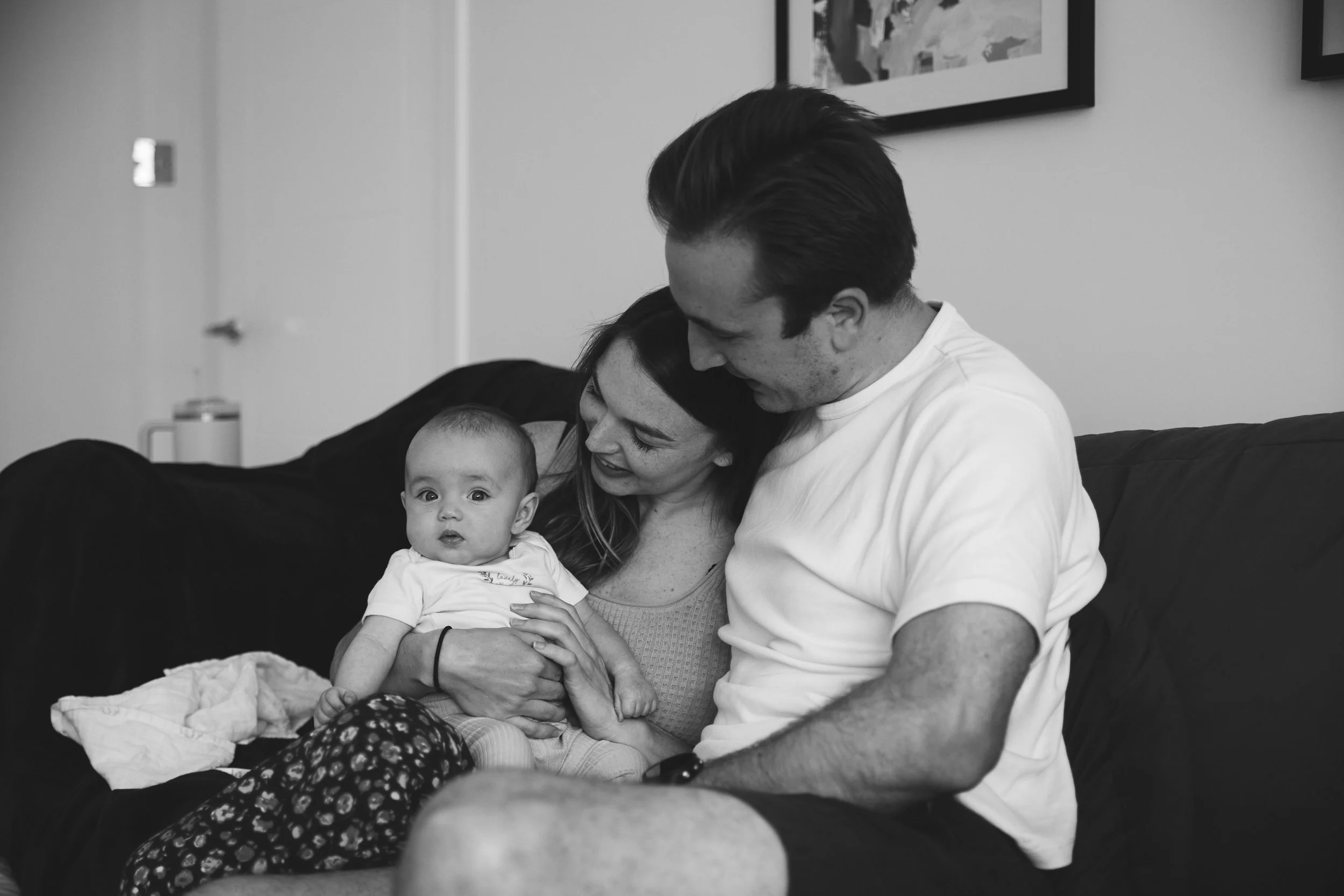 A happy family sitting on a couch, holding their baby and smiling at each other, in a living room.