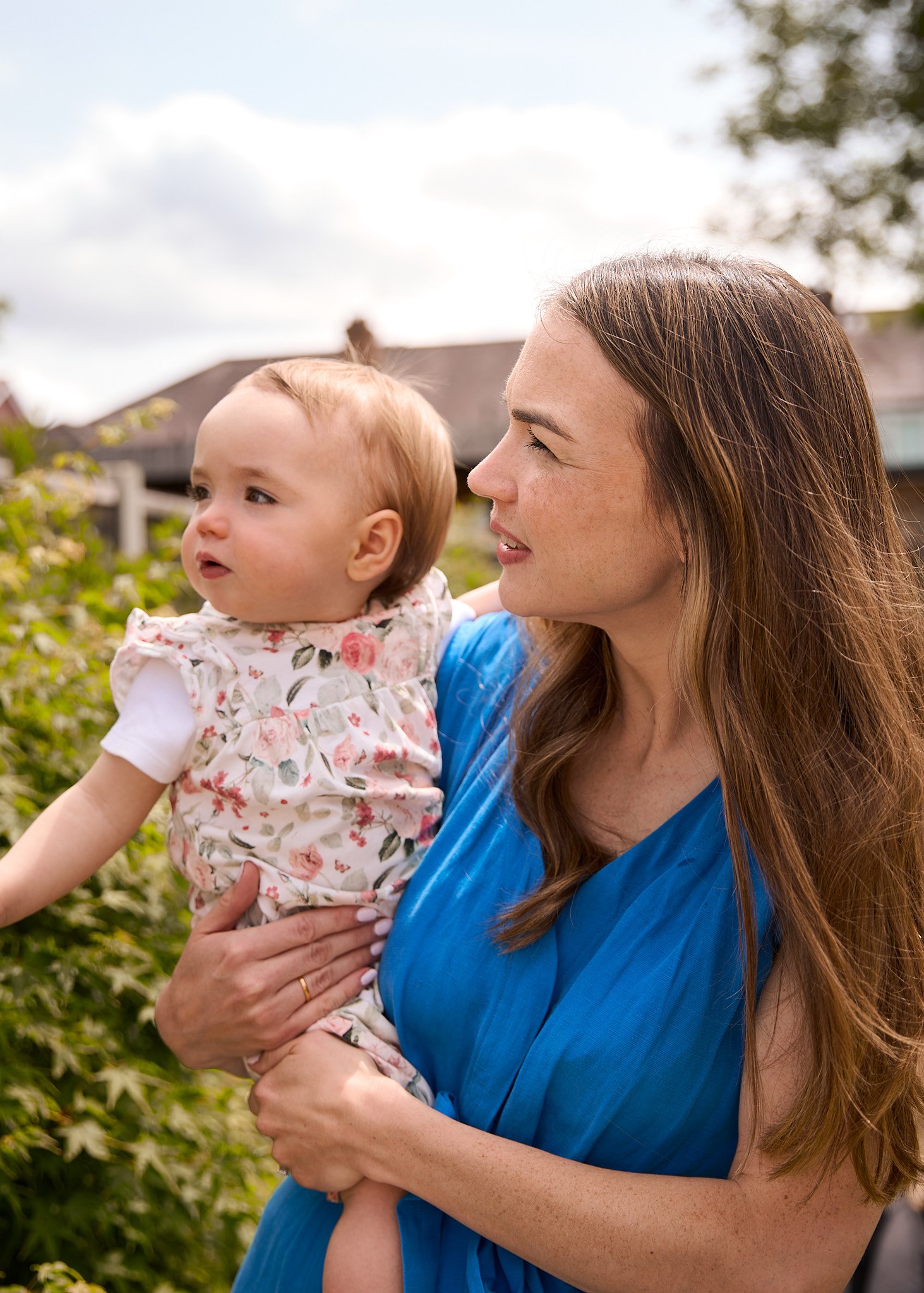 A woman holding a young girl outdoors in a garden with houses and cloudy sky in the background.