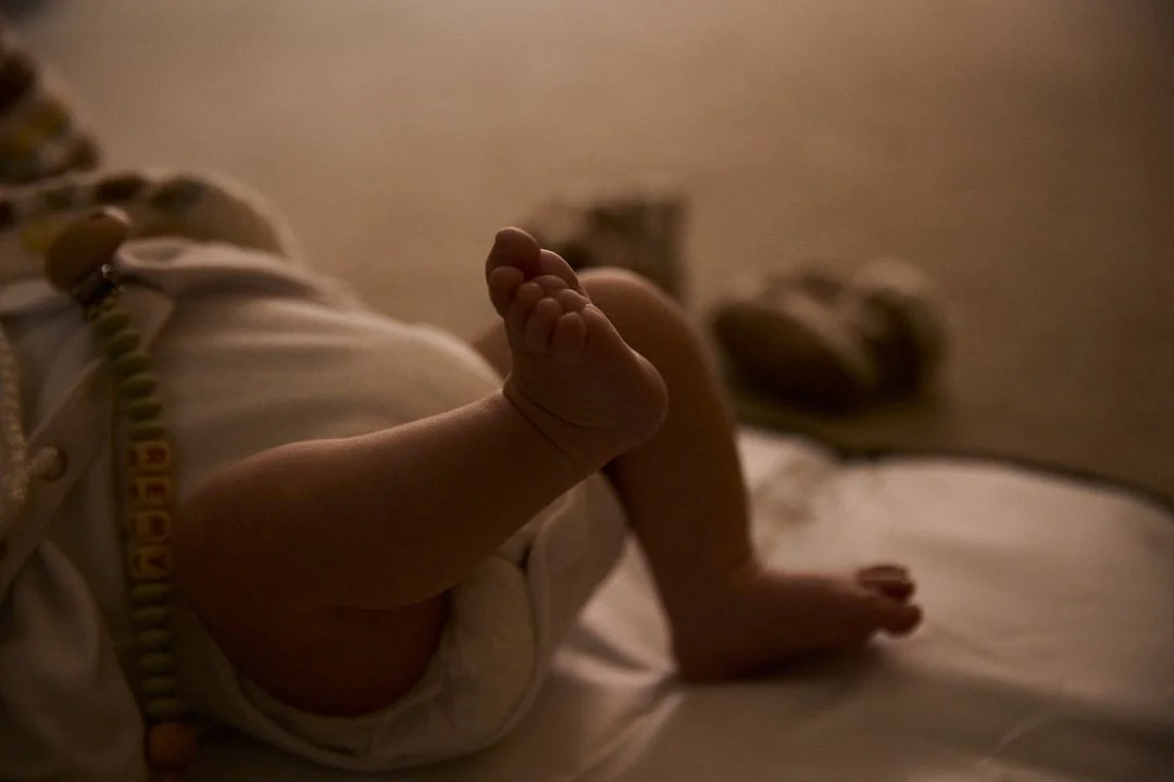 A baby lying on a bed, with one leg bent and foot raised in the air, wearing a white onesie and a pacifier clip. The room is dimly lit.