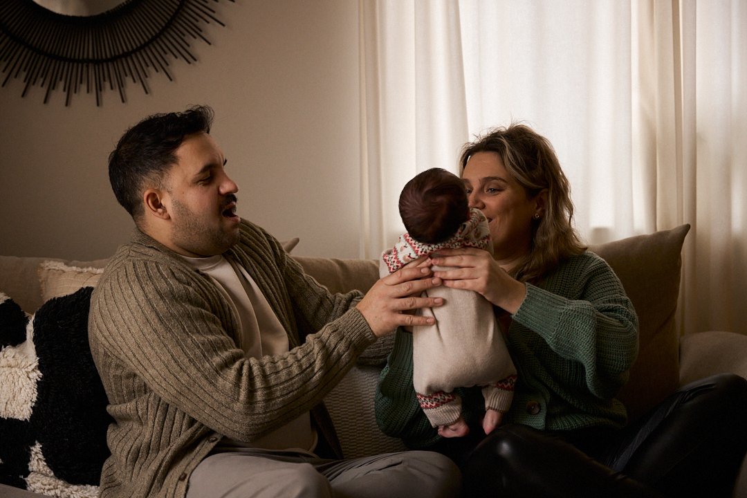 A family of three sitting on a couch, with a man and woman holding a young child between them, smiling and interacting indoors near a window with curtains.