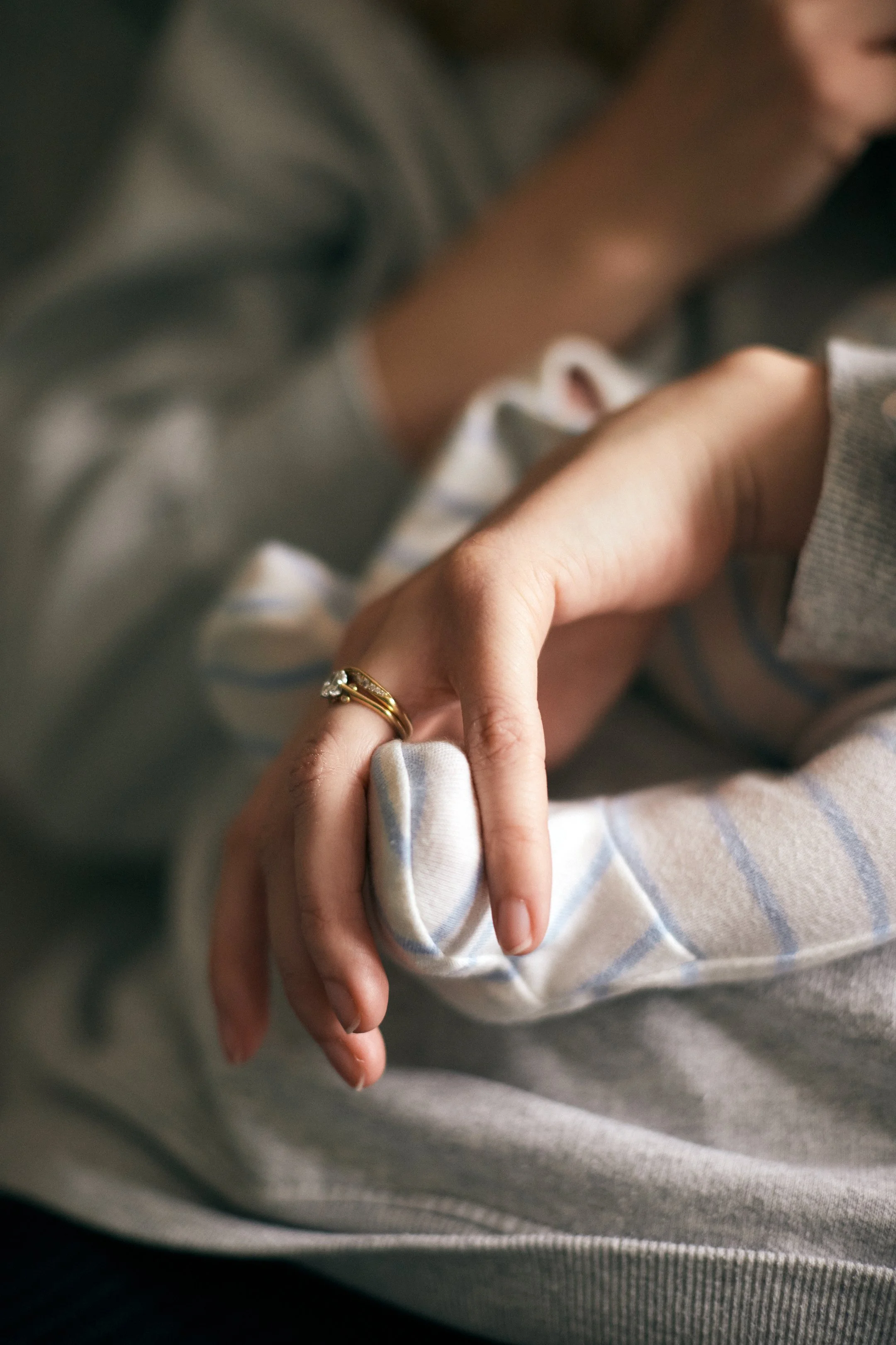 Close-up of an adult's hand gently holding a baby's hand, showing a gold ring with a gemstone on the adult's finger. The background is blurred, focusing on the hands.