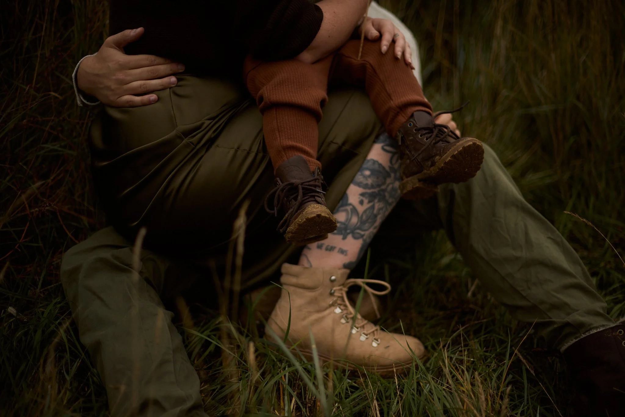 A child sitting on an adult's lap, with both wearing rugged outfits and boots, in the grass outdoors.