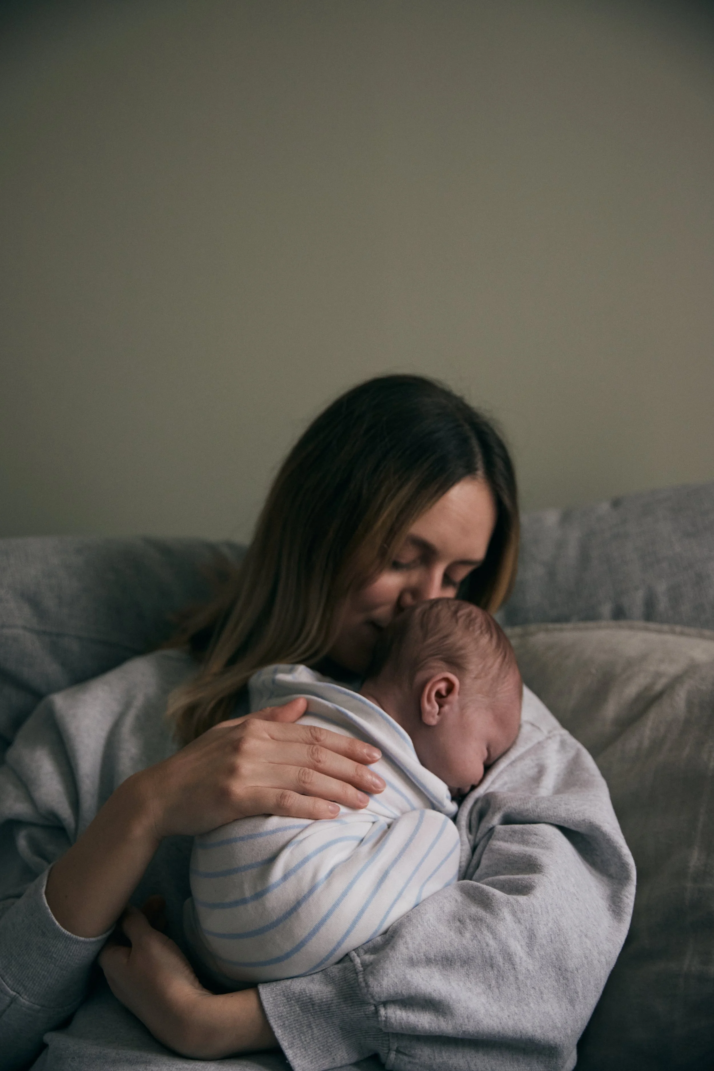 A woman with brown hair holding a sleeping baby dressed in a striped onesie close to her chest, sitting on a gray couch in a dimly lit room.