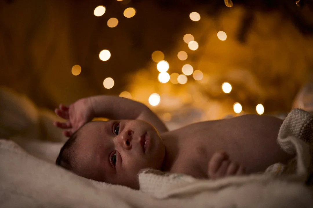 A baby lying on a blanket, looking at the camera, with warm blurred lights in the background.