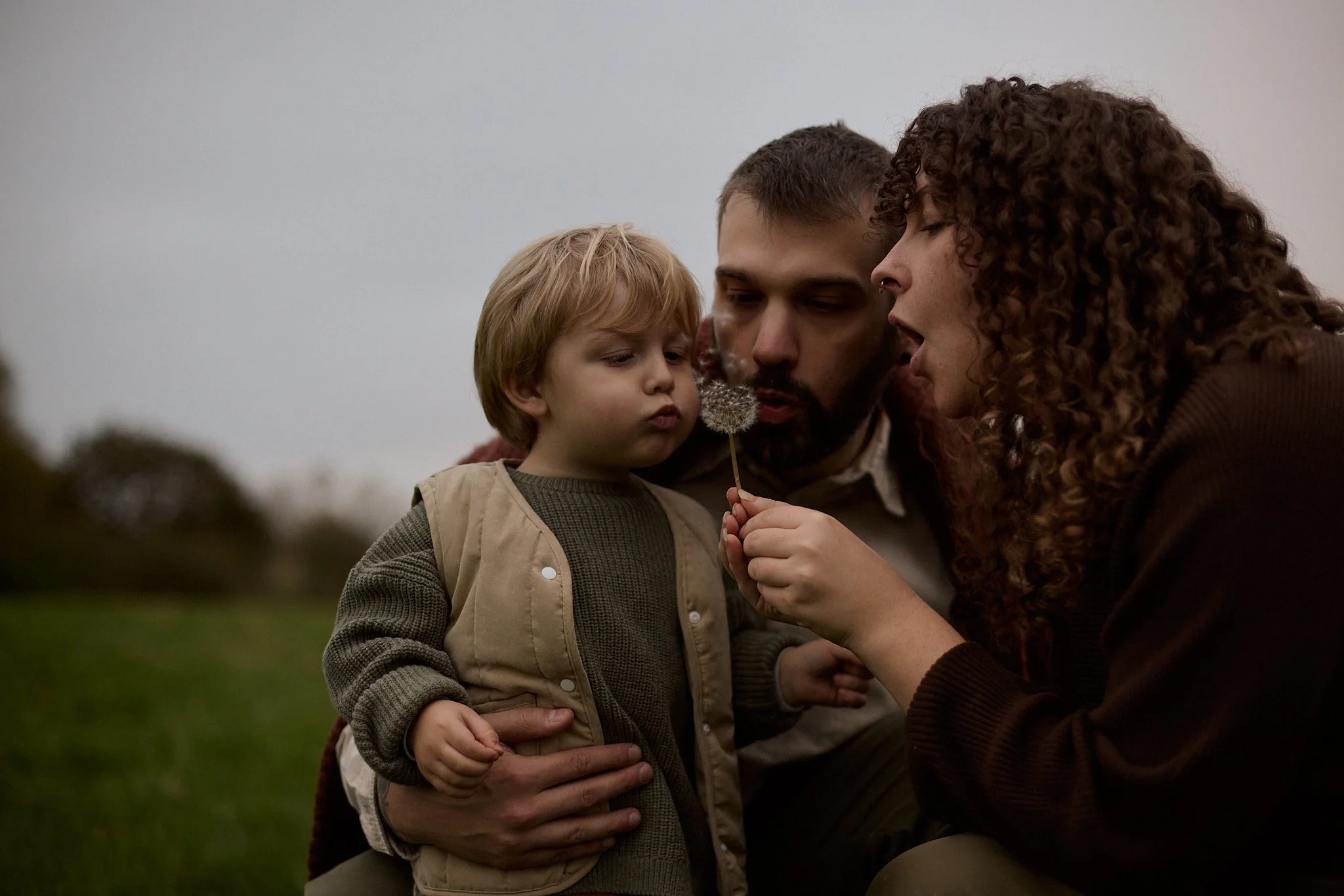 A family of three, a man, a woman, and a young boy, outdoors on a cloudy day, closely observing a dandelion seed head.