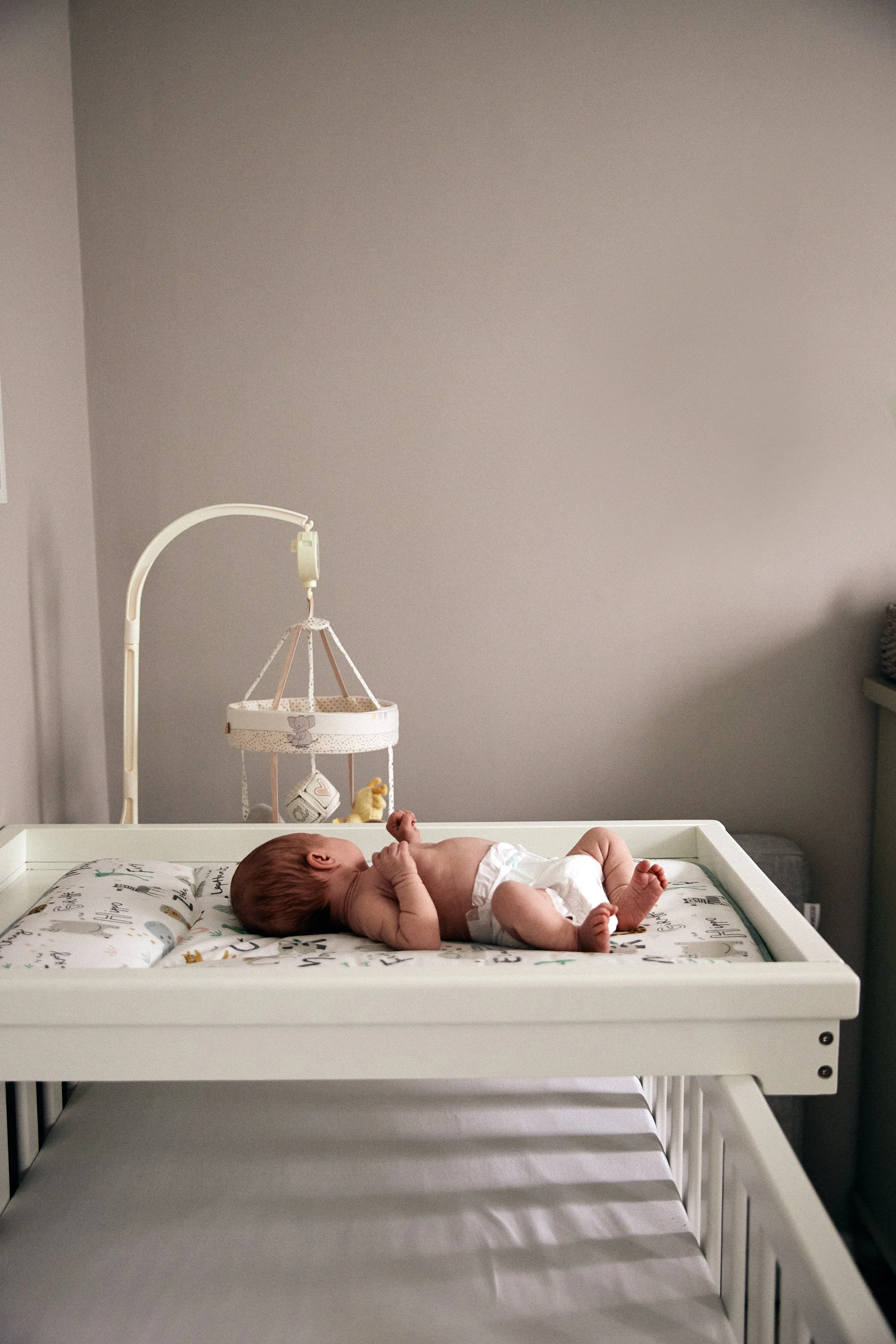 A newborn baby lying on its back on a changing table with a patterned sheet, surrounded by neutral-colored walls and furniture, with a crib underneath and a baby mobile hanging above.