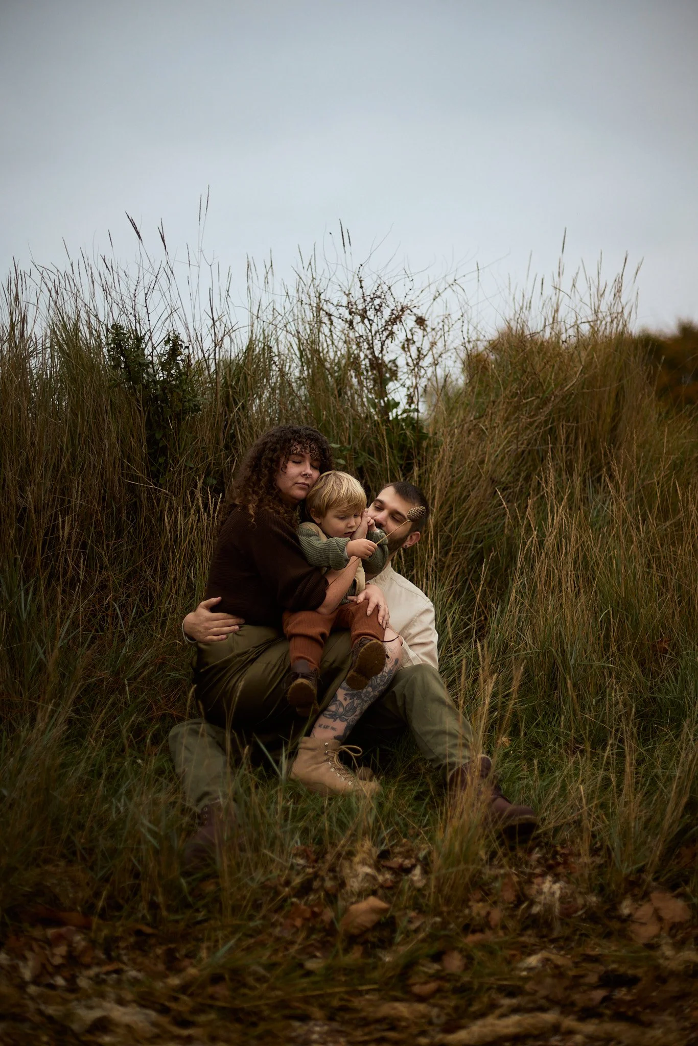 A family of three sitting outdoors among tall grass and dried leaves on a cloudy day. The woman is holding a young boy, and a man is sitting beside them, all appearing relaxed and enjoying each other's company.