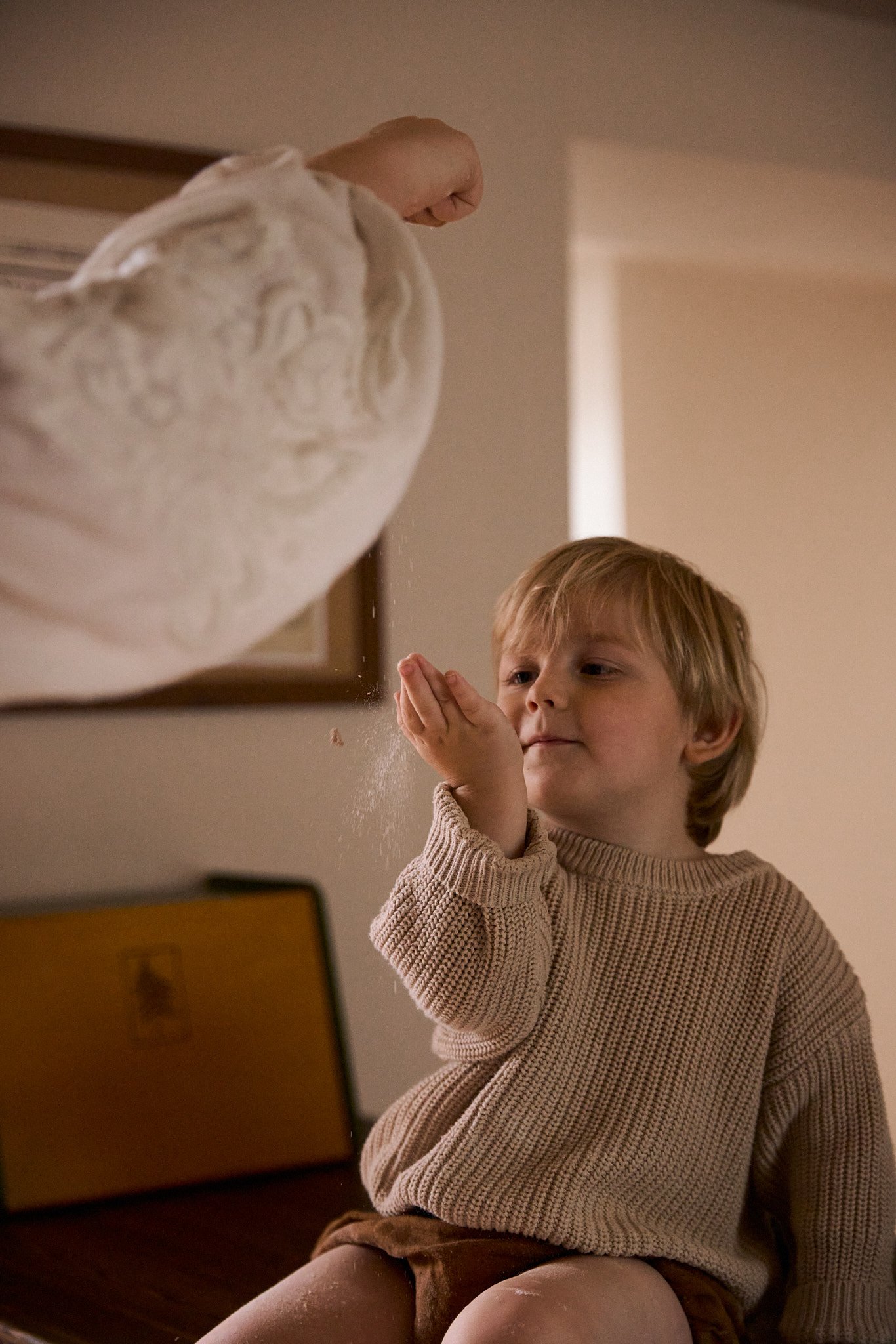 A young boy sits on a chair, examining a sculpture of a head with curiosity. An arm is seen tossing powder onto the sculpture's face.
