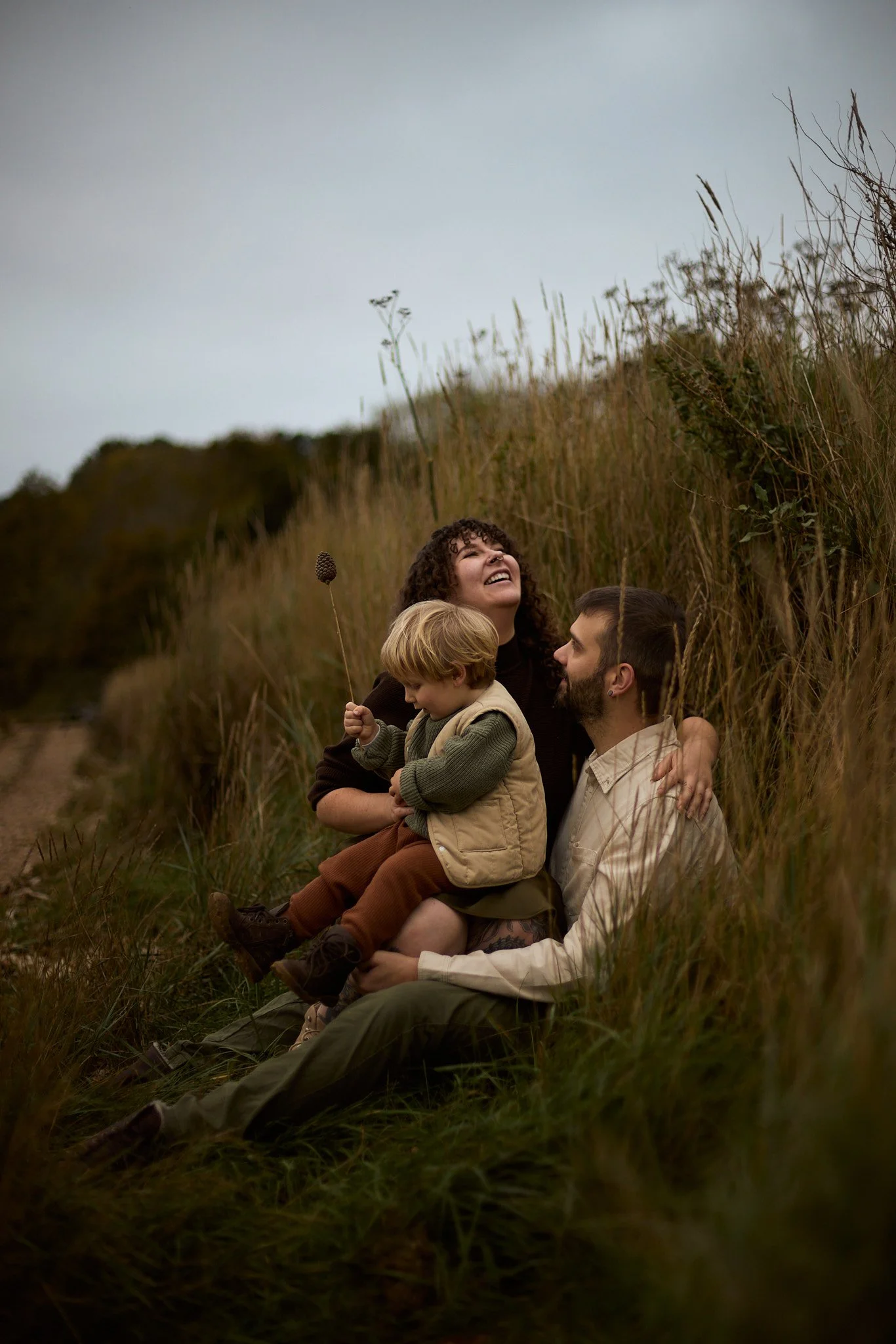 Family sitting outdoors amid tall grass, smiling and enjoying each other's company on a cloudy day.
