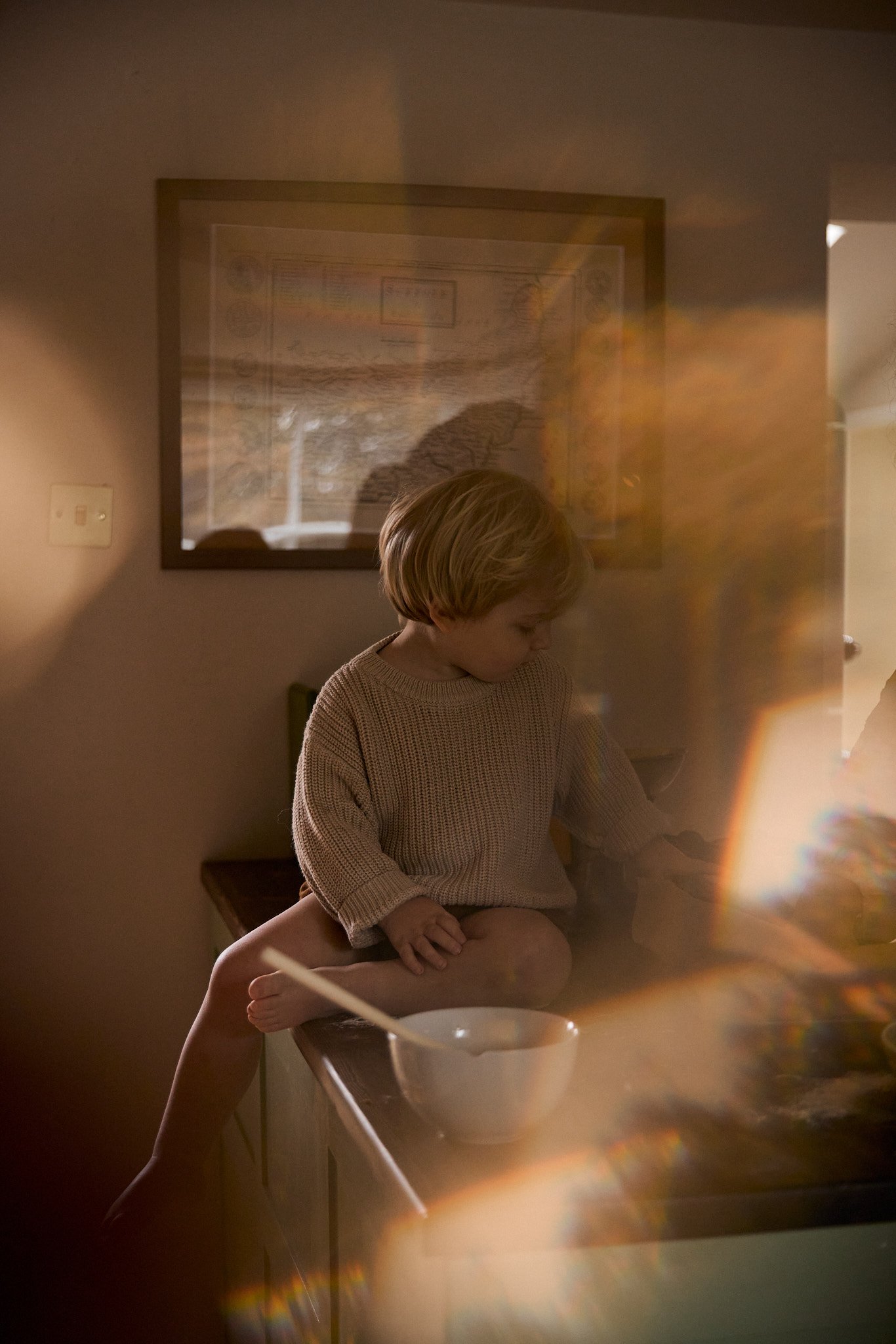 A young boy with blonde hair sitting on a kitchen counter, looking down, wearing a beige sweater. A bowl and spoon are in front of him. The photo has warm, soft lighting and is slightly blurry with a rainbow light reflection.