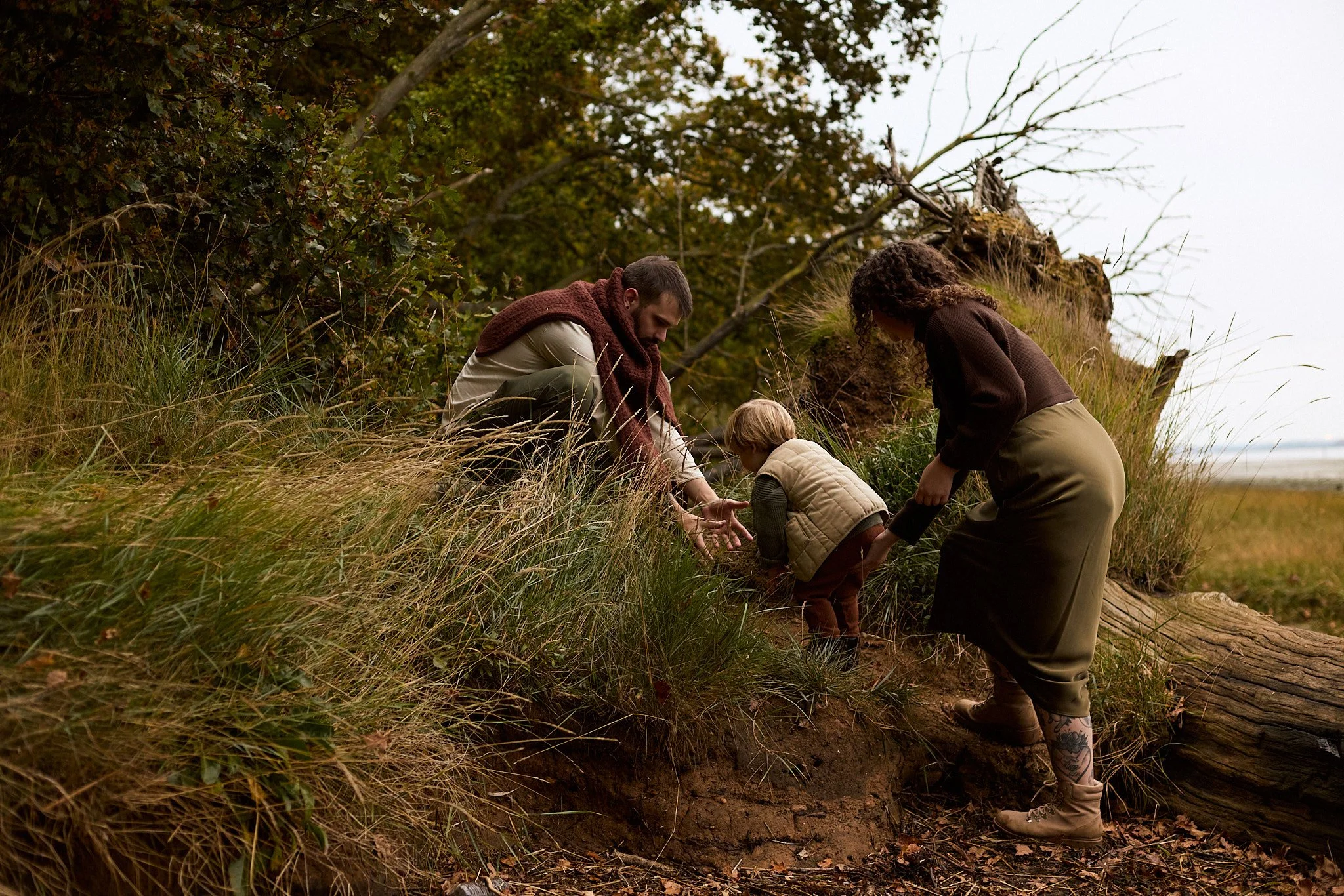 A family of three, two adults and a child, digging in the dirt outdoors amidst tall grass and trees, likely planting or exploring nature.
