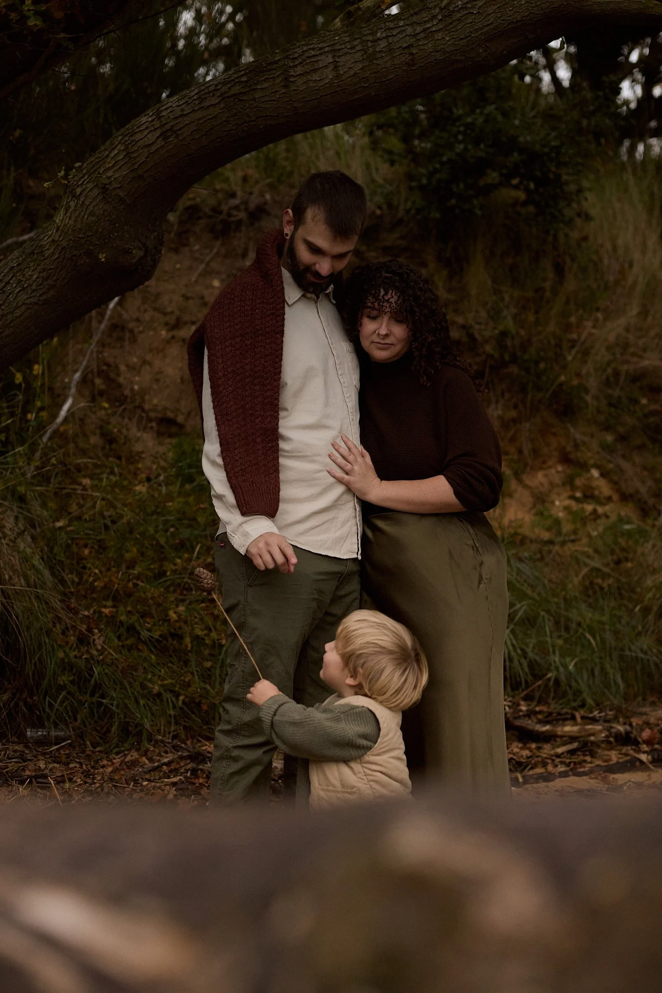 A family of three standing outdoors under a large tree, with the father and mother gently embracing and their young son holding a stick, in a natural, wooded setting.