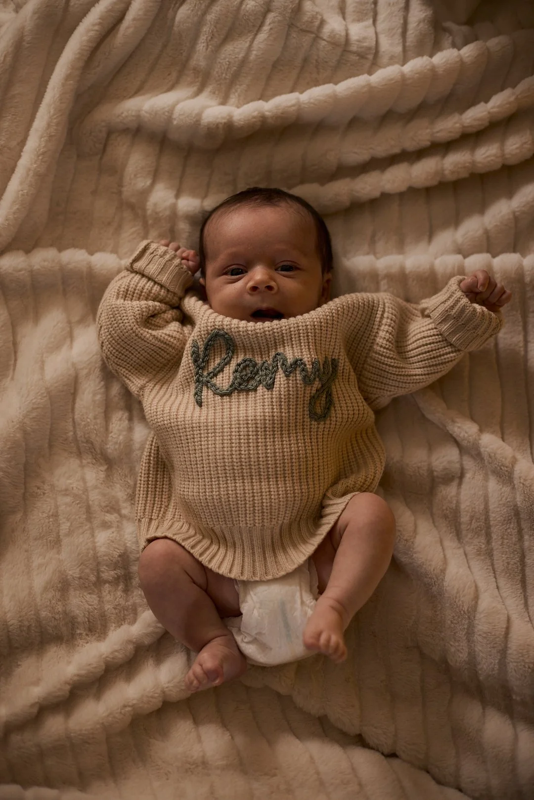 A baby lying on a soft, cream-colored blanket, wearing a beige sweater with the word 'Remmy' embroidered on it, raising fists in excitement or curiosity.