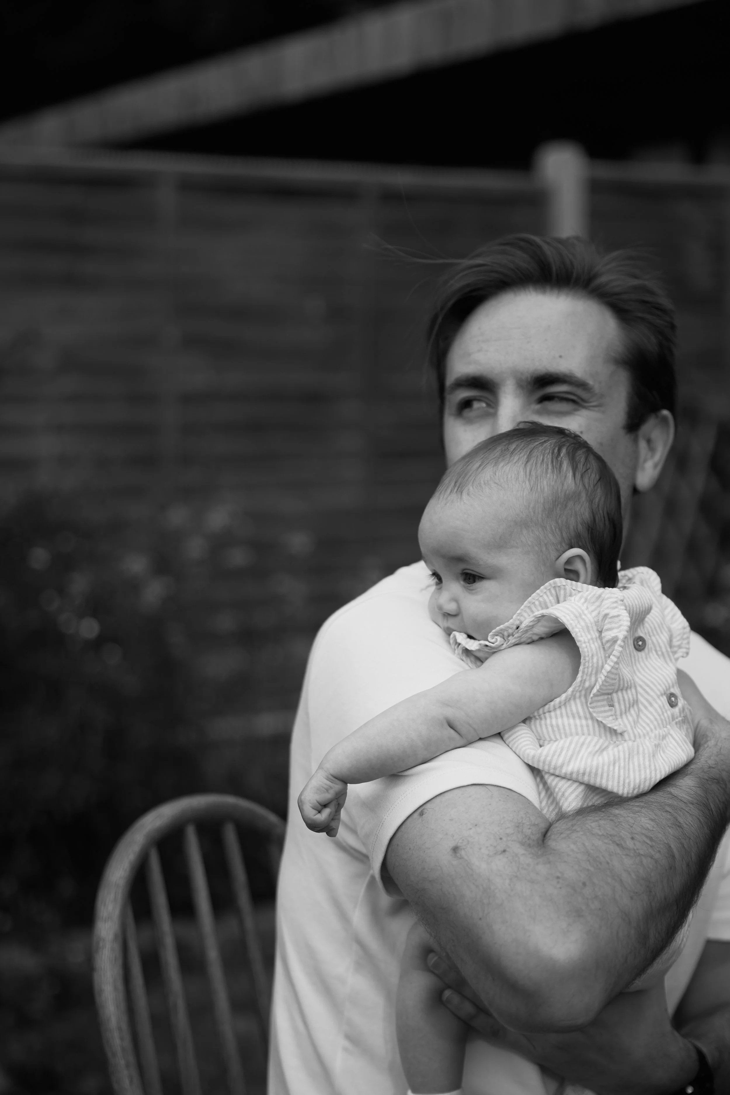A man holding a baby in his arms outdoors, with a wooden fence and a patio chair in the background, in black and white.