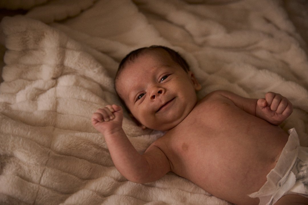 Smiling baby lying on a soft, cream-colored blanket, looking at the camera with arms slightly raised.