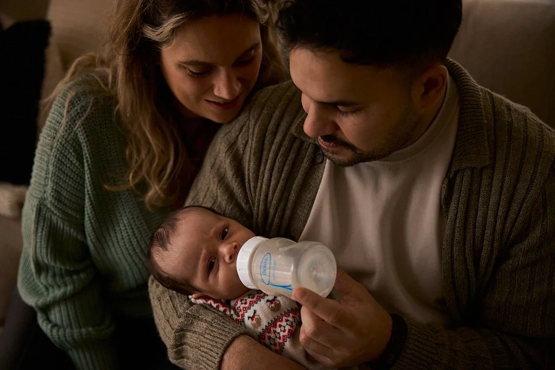 A young family—mother, father, and baby—gather around the baby, who is drinking from a bottle, in a cozy indoor setting.
