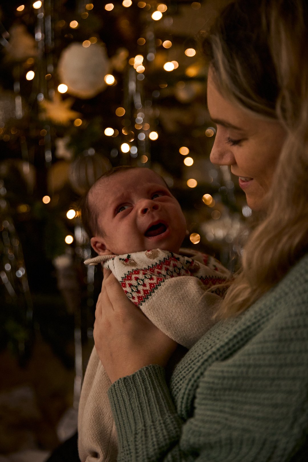 A woman holding a crying baby in front of a decorated Christmas tree with blurred lights.