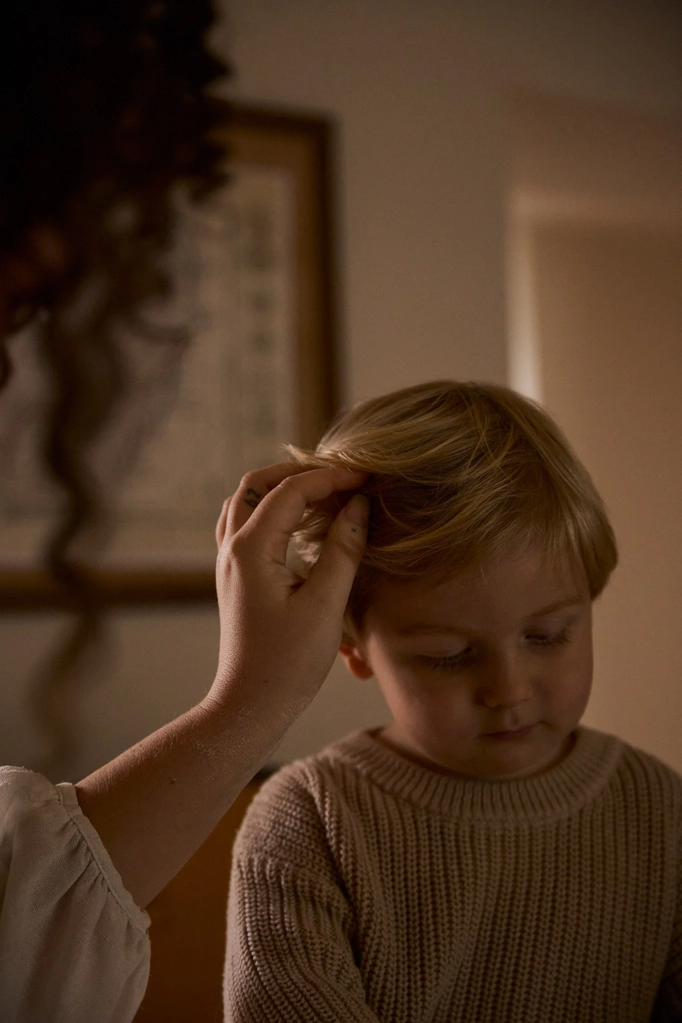 A person with curly hair is gently touching the blond hair of a young boy with freckles and a beige knit sweater, who appears to be looking down in a dimly lit room.