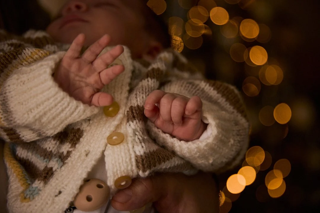 Close-up of a sleeping baby wearing a knitted sweater, with a blurred background of warm Christmas lights.