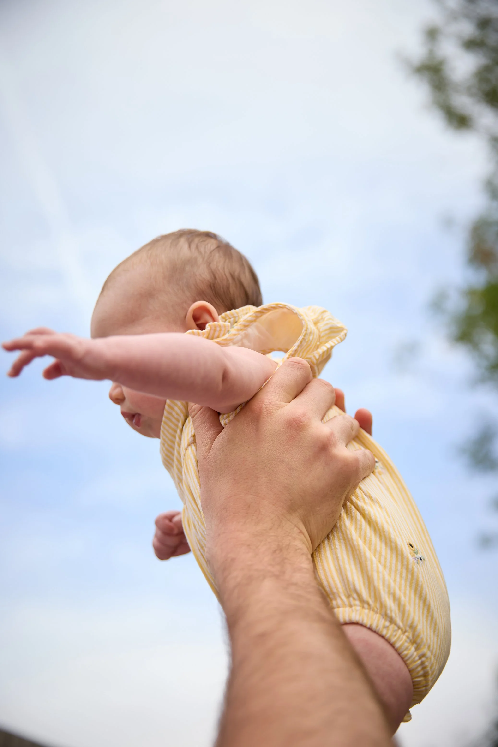 An adult holding a baby girl outdoors with her arm extended. The sky and trees are visible in the background.
