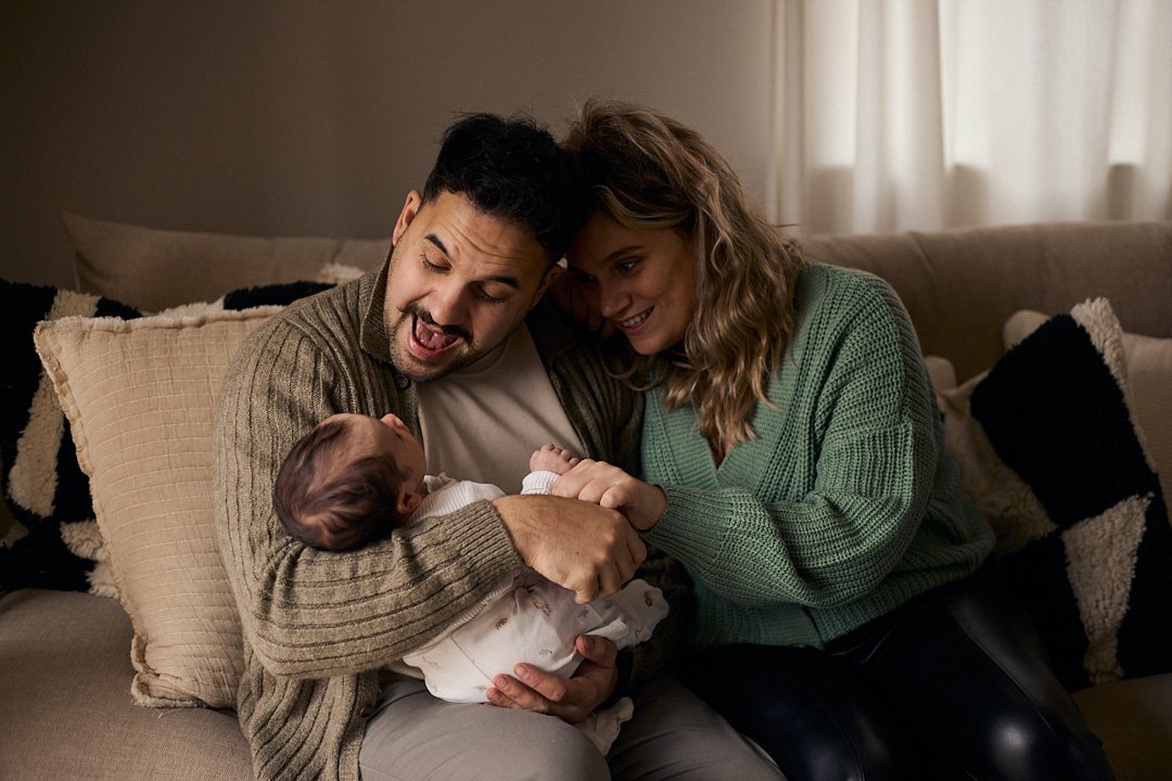 A family of three, including a man, a woman, and a baby, sitting on a beige couch with pillows. They are smiling and looking fondly at the baby, holding its hands gently.