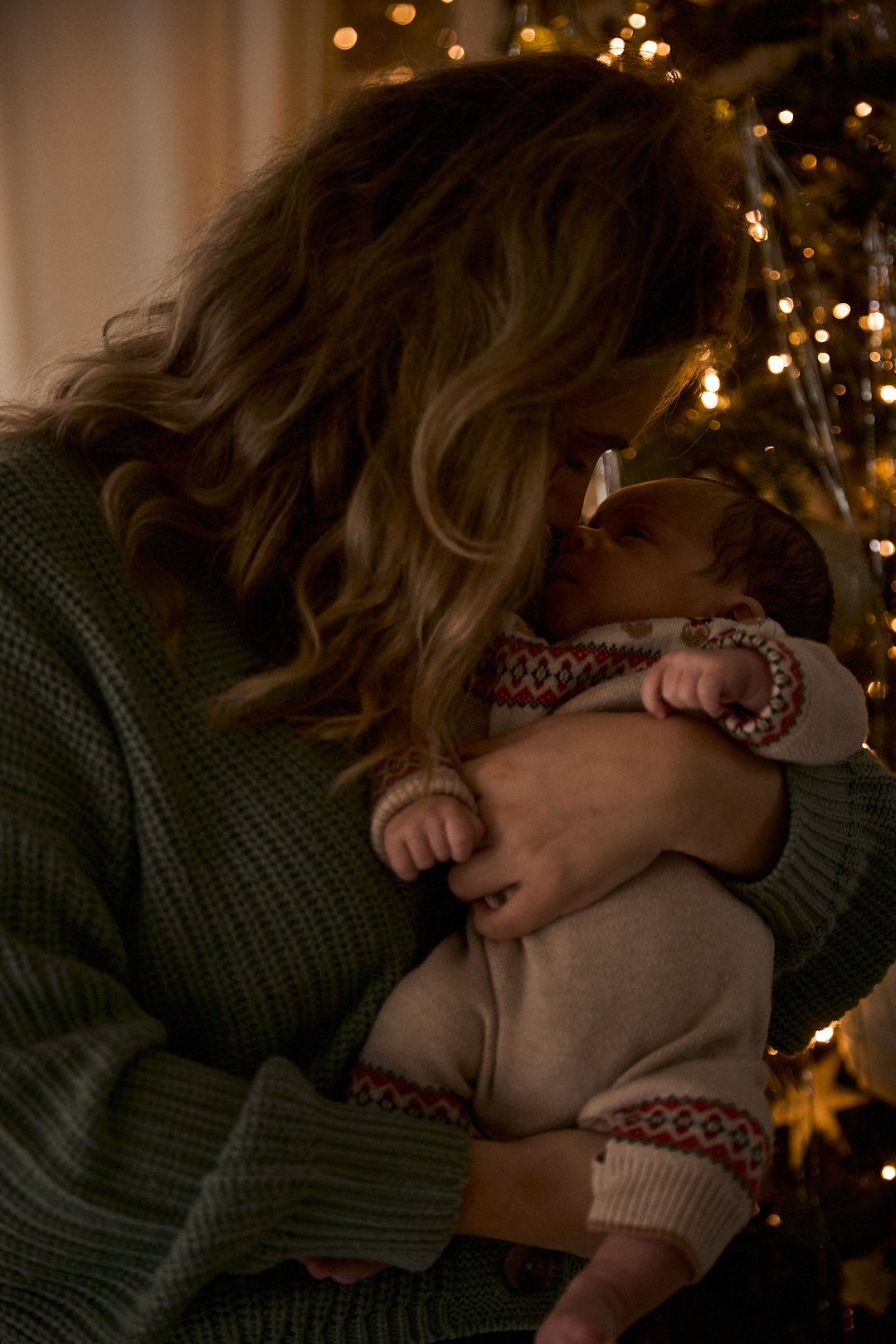 A woman holding a baby close, touching foreheads, with a Christmas tree decorated with lights in the background.