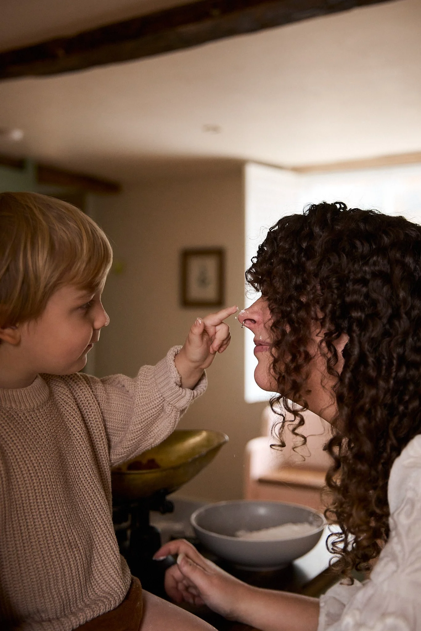 A young boy with light brown hair touching the nose of a woman with curly dark hair and a nose ring, in a cozy kitchen setting.