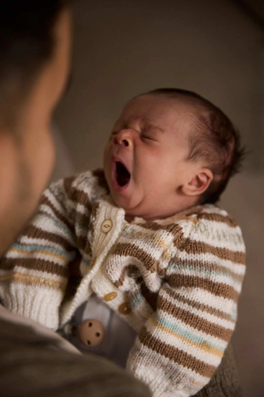 A yawning baby being held by an adult, seen from the side, with the baby's face expressive of tiredness or sleepiness.