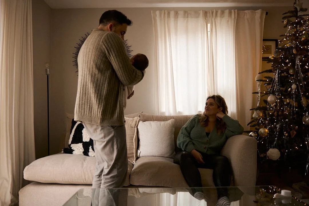 A man holding a baby in a cozy living room with a Christmas tree, a woman sitting on a sofa looking at them, warm lighting, with curtains and holiday decorations.