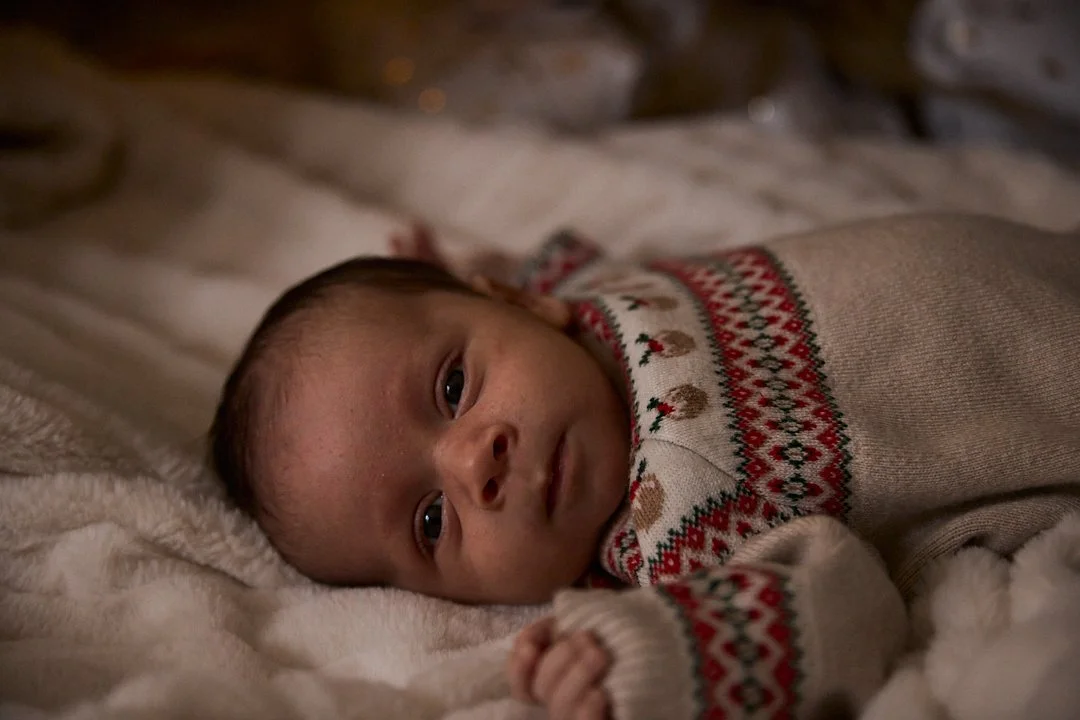 A baby lying on a soft blanket, wearing a festive sweater with holiday patterns, looking directly at the camera.