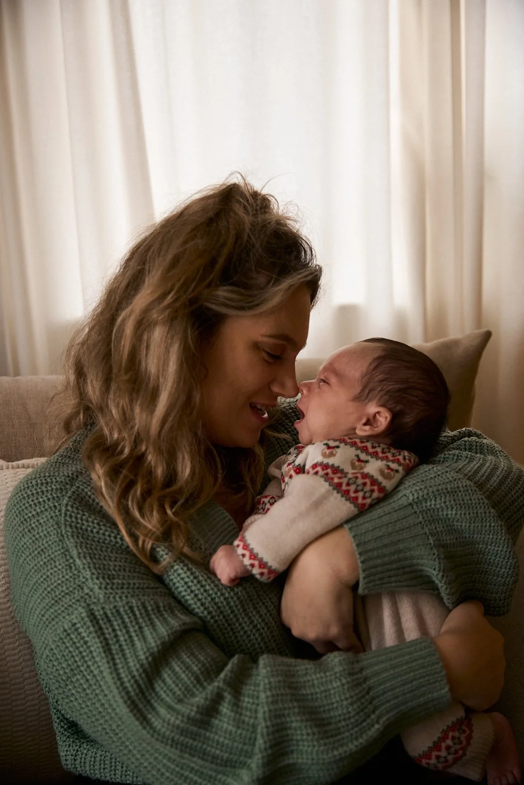 A woman holding a crying baby, both smiling and touching noses, in a cozy living room with light-colored curtains.