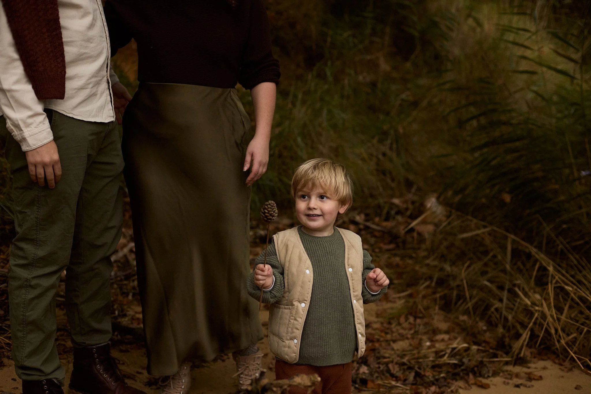 A young boy holding a pine cone on a stick stands outside on a trail, looking up and smiling, with two adults partially visible beside him. The scene is surrounded by tall grass and fallen leaves.