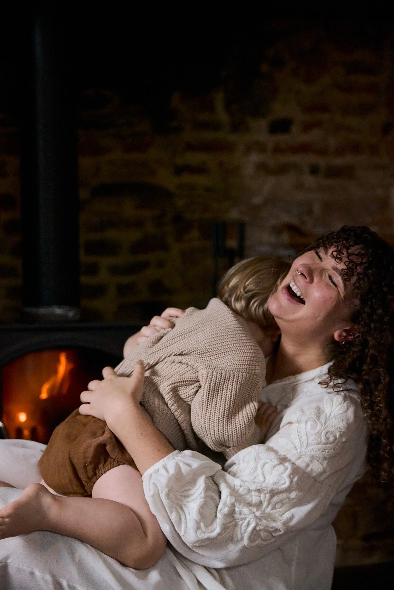 A woman with curly hair and earrings laughing and hugging a child in front of a fireplace.