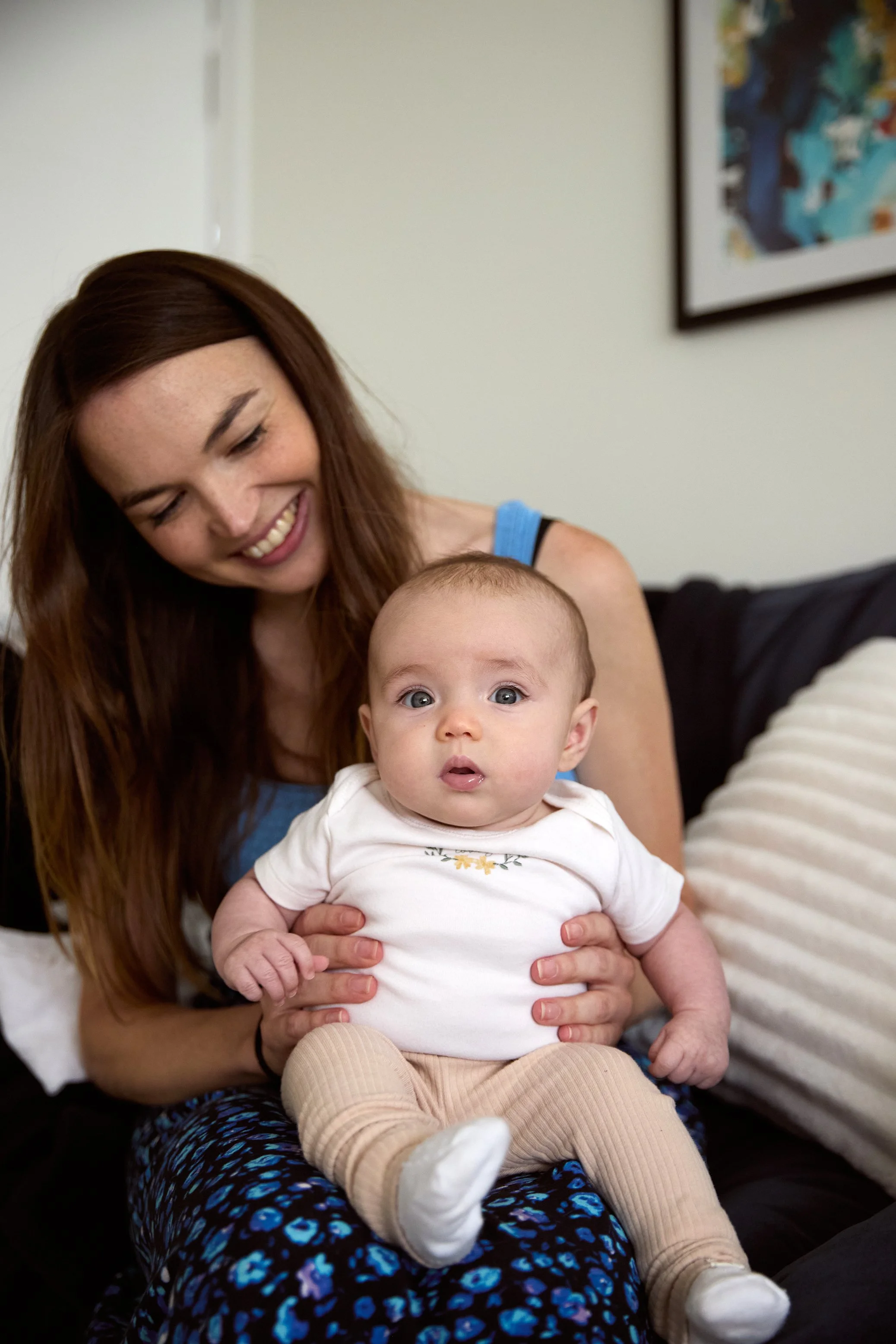 A woman with long brown hair smiling and holding a baby with blue eyes, wearing a white shirt and beige pants, sitting on a black couch in a living room.