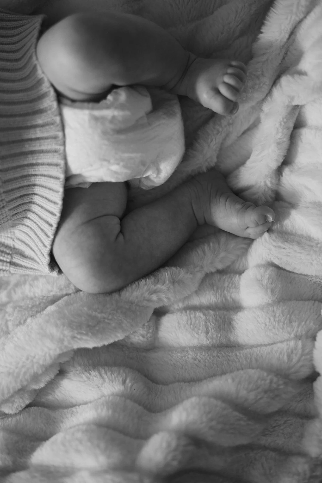 Close-up of a newborn baby's tiny feet resting on a soft, fluffy blanket, with part of the baby's legs and a piece of striped clothing visible.