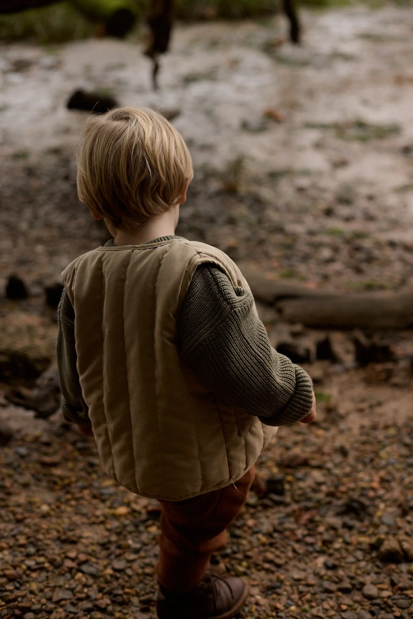 Back view of young child with blond hair, wearing a beige vest and brown pants, standing outdoors on a rocky path.