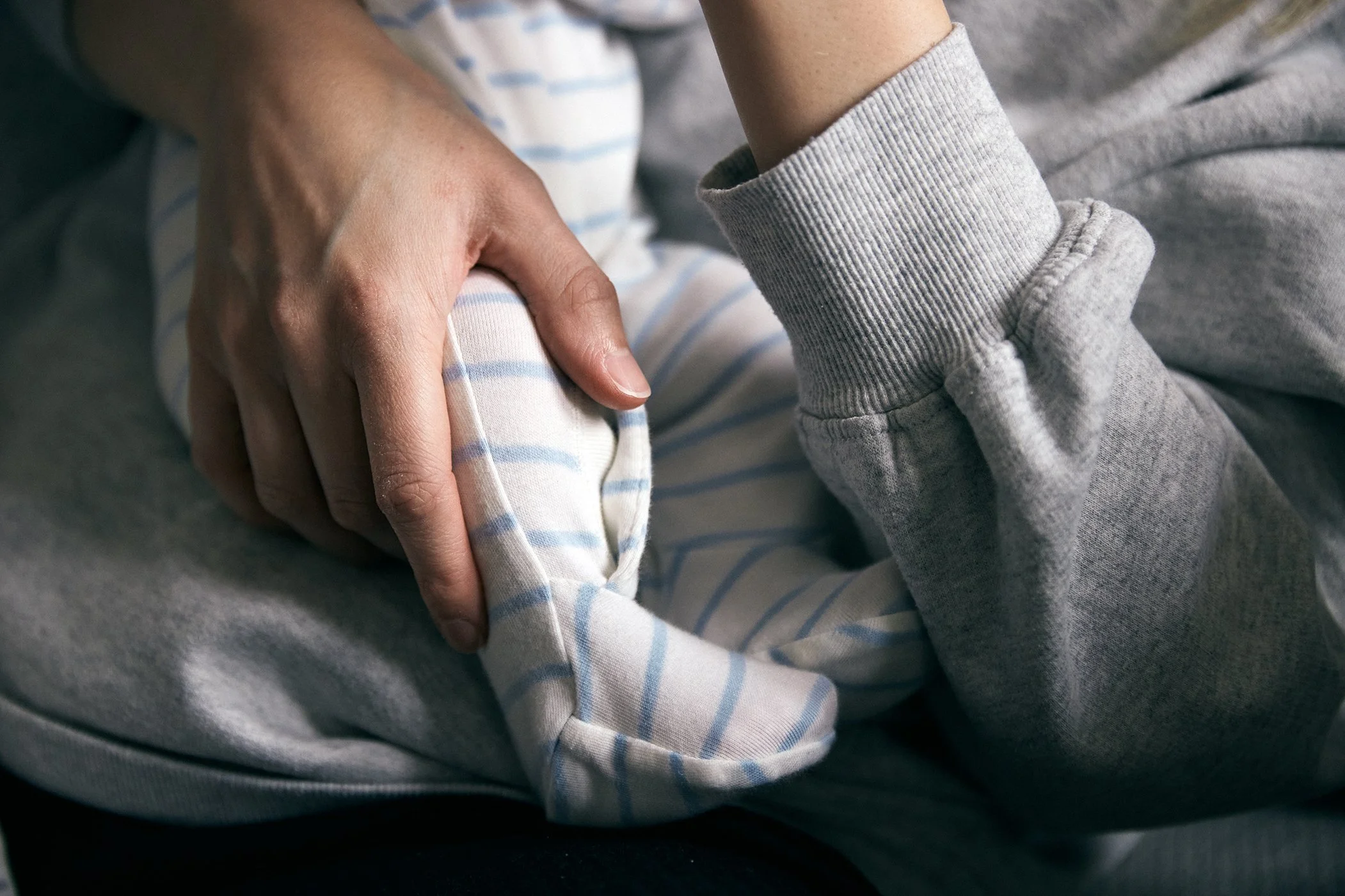 An adult holding a baby's foot gently, with the baby wearing striped pajamas and gray sweatshirt.