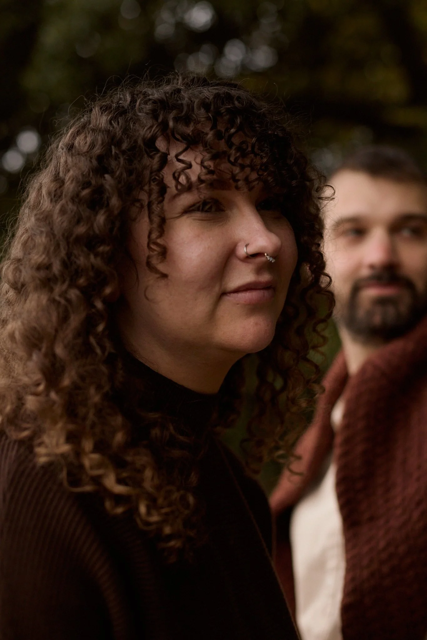 Close-up of a woman with curly hair and multiple facial piercings, looking to the side, with a man with a beard blurred in the background.