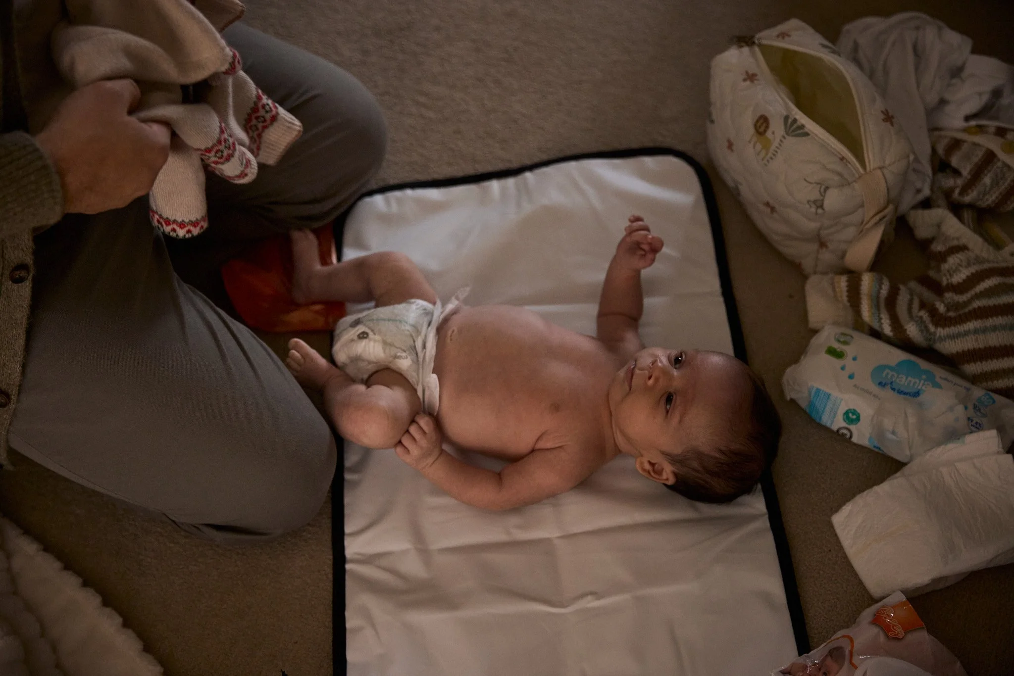 A baby lying on a changing pad on the floor, being attended to by someone holding a diaper.